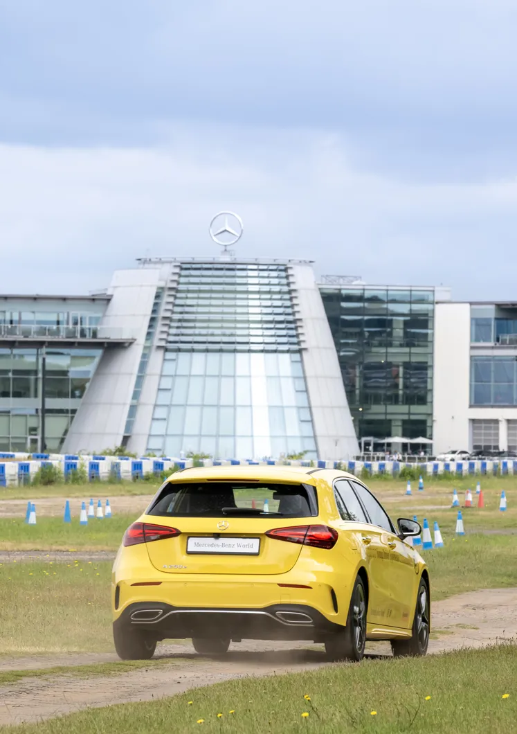 A bright yellow Mercedes-Benz A-Class drives on a track toward the modern glass entrance of Mercedes-Benz World, marked by the iconic Mercedes emblem on top.